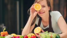 Bir kadın, yüzünün önünde bir portakal dilimi tutuyor. Masa üzerinde ise çeşitli sebze ve meyveler bulunuyor.