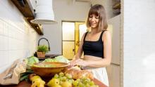 Kadın, üzerinde ahşap bir kase duran mutfak tezgahında armut, üzüm, marul ve ekmek ile yiyecek hazırlarken görülüyor.